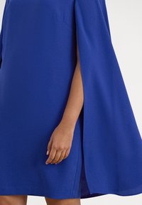 Modra obleka z ohlapnim krojem, ogrnjenimi rokavi in stranskim razporkom, izdelana iz gladkega materiala. Preprost dizajn brez vzorcev ali okraskov.