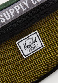 Close-up van een zwarte mesh zak met een gele stoffen achtergrond en een wit Herschel Supply Co. merklabel, naast olijfgroene stof met witte tekst.