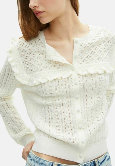 Donna che indossa un cardigan bianco a maglia con bottoni, caratterizzato da motivi a diamante e foglia, e dettagli arricciati su petto e spalle.