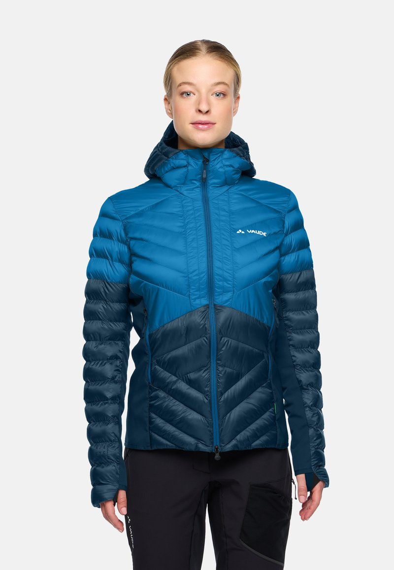 Donna che indossa una giacca a vento isolante di colore blu e verde teal scuro con zipper frontale e pantaloni neri, in piedi su uno sfondo chiaro.
