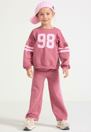 Vaaleanpunainen collegepaita, jossa on valkoinen numero 98 ja raidalliset hihat, yhdistetty samansävyisiin vaaleanpunaisiin housuihin. Vaaleanpunainen lippalakki on asetettu hieman vinoon.