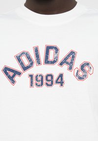 Λευκό βαμβακερό μπλουζάκι με φθαρμένο μπλε λογότυπο "ADIDAS" και το "1994" με κόκκινο περίγραμμα, με στρογγυλή λαιμόκοψη.