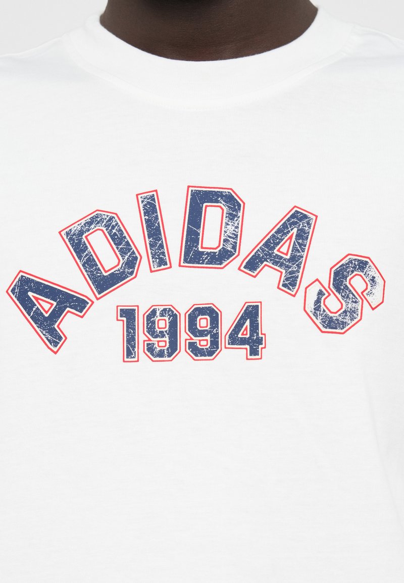 T-shirt en coton blanc avec un logo "ADIDAS" bleu usé et "1994" en contour rouge, doté d'un col rond.