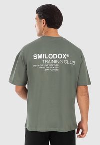 Camiseta verde oliva de manga corta y cuello redondo. Presenta texto en blanco en la parte posterior: "SMILODOX TRAINING CLUB."