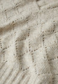 Pletená tkanina v béžové barvě s diamantovým vzorem. Měkká textura a žebrované okraje, s malými perforacemi po celém designu.