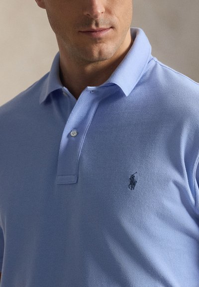 Άνδρας που φοράει ένα ανοιχτό γαλάζιο polo με γιακά και δύο κουμπιά, με μικρό σκούρο κεντημένο λογότυπο στο αριστερό στήθος.