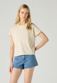 Vaaleanbeige puuvillapaidan, jossa on lyhyet hihat, etupuolella on kohokuvioitu "LEVI'S" -logo. Se on yhdistetty sinisiin farkkushortseihin.