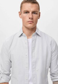 Camisa a rayas de color gris claro con cuello abotonado, mangas enrolladas y una camiseta blanca visible debajo. Tela texturizada con rayas verticales.