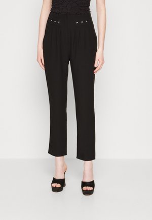 Kobieta ubrana w czarne, proste spodnie z wysokim stanem i detalami w postaci ćwieków, w połączeniu z czarnymi, platformowymi sandałami na otwartym palcu, stojąca na białej podłodze.