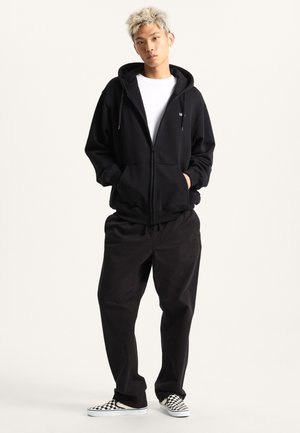 Czarna bluza z kapturem na zamek błyskawiczny i ze ściągaczem, noszona na białej koszulce. Połączona z luźnymi czarnymi spodniami i wkręcanymi butami w kratę.