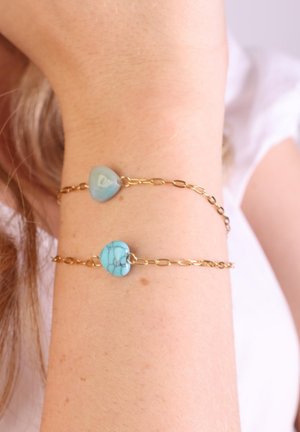 Gouden kettingarmband met twee hartvormige edelstenen: een lichtblauw en een turkoois. Gladde textuur, de schakels zijn licht verlengd.