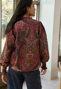Multicolored, patterned blouse with long puffed sleeves, ribbed texture, and a high neck, paired with dark denim pants.