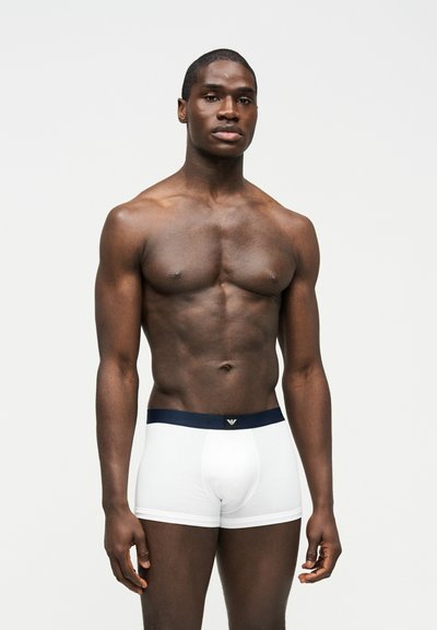 Μυώδης άνδρας με σκούρο δέρμα στέκεται μπροστά σε απλό ανοιχτόχρωμο φόντο, φορώντας λευκά μπόξερ με σκοτεινή μέση.