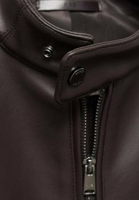 Veste en cuir brun foncé avec un col haut, dotée d'une fermeture éclair argentée et d'accents de boutons. Texture lisse, design minimaliste.