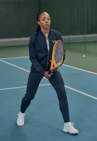 Čierna športová bunda a legíny, biele tenisky, drží oranžovú tenisovú raketu na modrom kurte s pozadím zelenej siete.