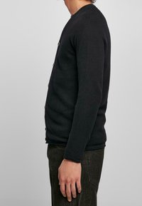 Svart texturerad kofta med rund halsringning och knäppning, bära över mörka byxor, visad från sidan. Enkel och funktionell design.