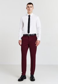 Camicia bianca a maniche lunghe, cravatta nera, pantaloni bordeaux, cintura nera e scarpe nere. Vestibilità slim con una texture liscia, accenti minimi.