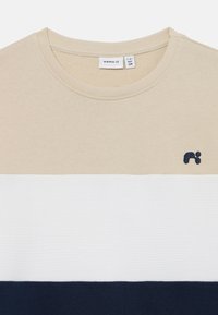 Beige og hvit bomulls t-skjorte med marineblå kant, med en teksturert hvit stripe og en liten brodert logo på brystet.