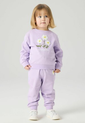 Småbarn med blondt hår iført en lavendel sweatshirt og bukser med hvite blomstermønstre og hvite joggesko, stående på en ensfarget bakgrunn.