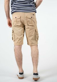 Shorts cargo beiges avec plusieurs poches latérales et arrière, une coupe décontractée et un détail de cordon aux ourlets, associés à des chaussures grises et noires.