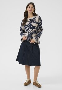 Žena stojaca v pulóvri s béžovo-tmavomodrým vzorom, tmavej rifľovej sukni po kolená, v hnedej teniske a s veľkými kruhovými náušnicami na bielom pozadí.