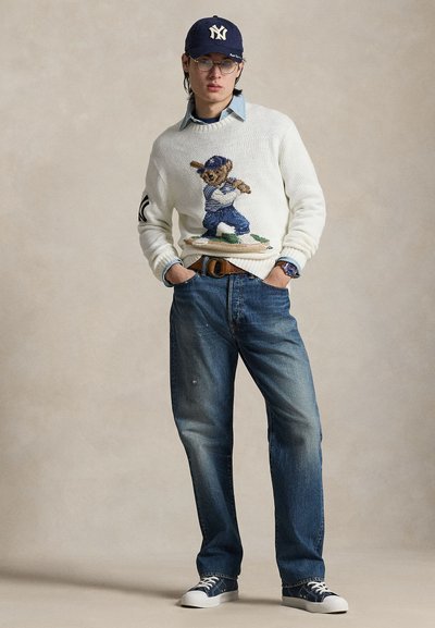 Λευκό πλεκτό πουλόβερ με σχέδιο αρκούδας σε στολή μπέιζμπολ, συνδυασμένο με μπλε τζιν και ναυτικά παπούτσια, ολοκληρωμένο με ναυτικό καπέλο.