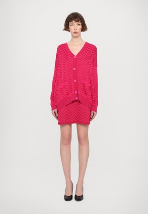 Cardigan fucsia con motivo zigzag, maniche lunghe, scollo profondo a V e bottoni, indossato sopra un vestito aderente abbinato, abbinato a tacchi neri.
