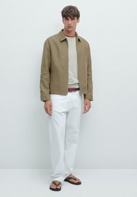 Giovane uomo che indossa una giacca beige, un maglione chiaro, pantaloni bianchi e infradito nere, appoggiato a un muro bianco semplice.