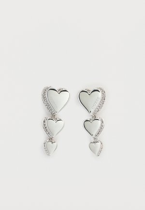 Boucles d'oreilles en forme de cœur argentées avec trois cœurs en cascade, rehaussées de strass scintillants. Surface réfléchissante avec le nom de la marque gravé.