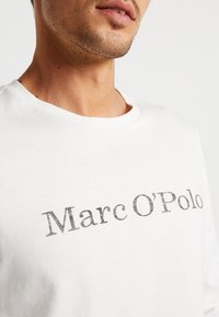 Witte katoenen t-shirt met een grijs, verouderd tekstontwerp dat "Marc O'Polo" leest, met een ronde halslijn en korte mouwen.