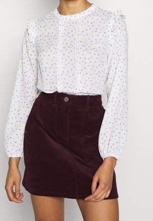 Vrouw draagt een witte blouse met lange mouwen met paarse en gele polkadots, gecombineerd met een hooggetailleerde donkerpaarse corduroy rok.
