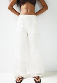 Weiße lässige Hosen aus Baumwolle mit vorderen Taschen und einem Kordelzugbund. Kombiniert mit braunen Flip-Flops. Einfaches Design.