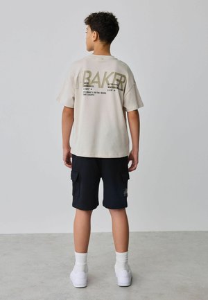 Teen boy stands facing away wearing beige graphic t-shirt, black cargo shorts, white socks, and white sneakers against plain background.