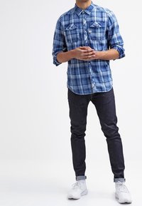 Man står med händerna sammanflätade, iklädd en uppvikt blårutig skjorta, mörka jeans och vita sneakers mot en enfärgad vit bakgrund.