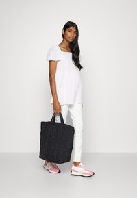 Bolsa tote acolchoada preta com padrão de ondas; usada com uma blusa branca texturizada, calças brancas e ténis com detalhes em rosa.
