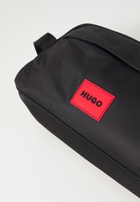 Čierny nylonový batoh so štvorcovým tvarom a výraznou červenou obdĺžnikovou značkou s nápisom "HUGO" v čiernom písme. Textúrovaný povrch.