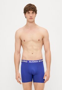 Zilas bokseršortus, izgatavotus no gludas auduma ar pieguļošu apgriezumu, ar kontrastējošu purpura krāsas jostu, uz kuras ir uzraksts "BJÖRN BORG".