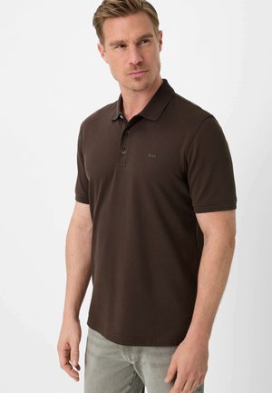 Uomo che indossa una polo a maniche corte di colore marrone scuro con colletto, in piedi davanti a uno sfondo chiaro e uniforme.