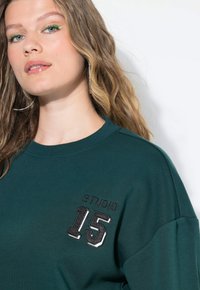 Joven con cabello rubio ondulado que lleva un sudadera verde con el texto "STUDIO 15" y delineador verde neón, posando contra un fondo blanco.