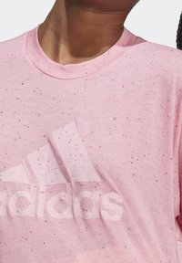 Vaaleanpunainen t-paita, jossa on pilkullinen kuvio, pyöreä kaula-aukko ja etupuolella näkyvä haalistunut Adidas-logo. Pehmeä kangasrakenne.