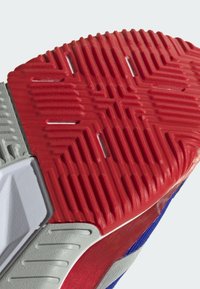 Semelle de chaussure de sport avec une surface en caoutchouc rouge texturé, présentant des accents gris et des rainures pour une meilleure adhérence. Léger éclat sur les bords.