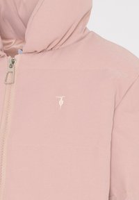 Veste à capuche rose clair avec zip, arborant un petit logo blanc et un tissu lisse et mat. Comprend des accents brodés et des détails de couture.