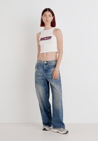 Hvitt kort tank-top med lilla tekst, kombinert med løst sittende lyseblå denimjeans. Modellen har på seg grå joggesko. Enkel bakgrunn.