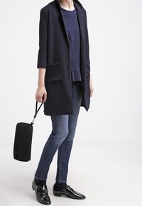 Manteau long bleu marine avec des manches 3/4, deux poches avant, porté sur un top peplum bleu marine et un jean, associé à des chaussures en cuir verni noir.