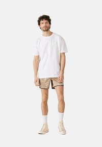 T-shirt bianca in cotone abbinata a pantaloni corti beige lucidi con profili neri, indossati con calzini bianchi alla caviglia e sneakers beige.