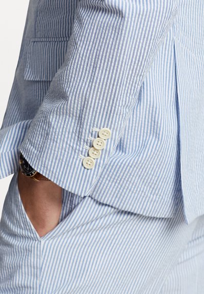 Tissu rayé bleu clair et blanc, poignet de blazer ajusté avec quatre boutons crème, mettant en valeur des coutures soignées et des détails de confection précis.