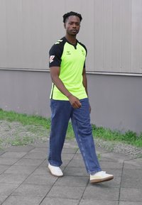 Maglia sportiva verde neon con maniche nere, abbinata a jeans in denim blu larghi e sneakers bianche. In piedi su una superficie pavimentata, con erba sullo sfondo.