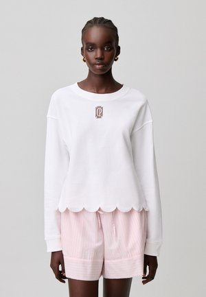 Giovane donna che indossa un top bianco a maniche lunghe con orlo smerlato e pantaloncini rosa a righe con coulisse su sfondo semplice.