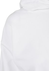 Bela jopa s kapuco z mehko teksturo, ki ima veliko kapuco in široke rokave. Tkanina je videti debela in rahlo obdelana, kar povečuje toploto.