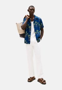 Camisa azul de manga corta con patrón de palmeras, top blanco, pantalones blancos y sandalias marrones. Lleva un bolso tote color beige y marino.